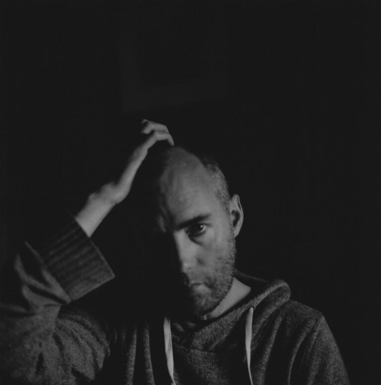 Black and white portrait of a man with a contemplative expression, hand on his head, in a dark setting.
